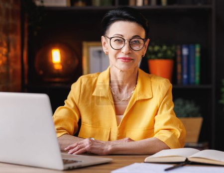 Photo for Trendy middle aged woman in yellow shirt and glasses smiling and  looking at camera  while sitting at table and resting during break in remote work in home office - Royalty Free Image