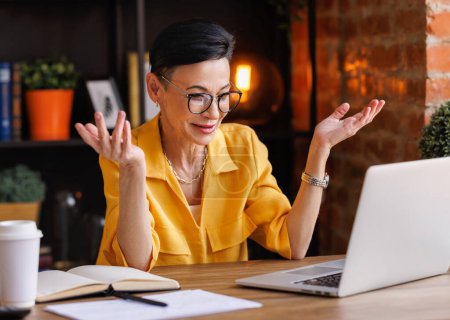 Photo for Glad middle aged female entrepreneur in stylish yellow blouse and glasses smiling and gesticulating while sitting at desk in home office and talking to online coworkers during online meeting - Royalty Free Image