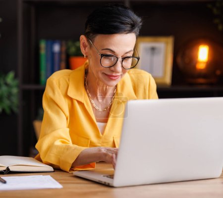 Téléchargez les photos : Femme manager d'âge moyen en chemisier élégant avec des lunettes tapant sur le clavier netbook tout en étant assis à la table et travaillant sur un projet d'entreprise dans le bureau à la maison avec des murs de briques - en image libre de droit