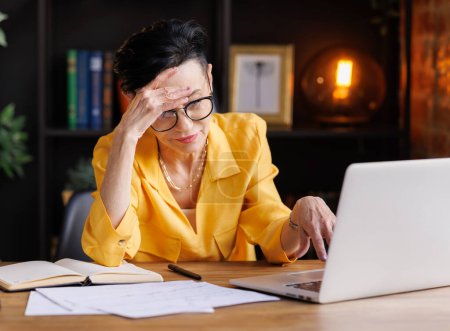 Photo for Tired mature female manager in trendy yellow blouse and glasses rubbing temples while sitting at table with papers and laptop and suffering from headache during work in home office - Royalty Free Image