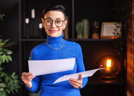 Foto de Autoasegurada mujer de mediana edad en cuello alto azul y gafas elegantes con documentos mirando a la cámara con sonrisa mientras está de pie cerca de la pared de ladrillo en la oficina en casa - Imagen libre de derechos