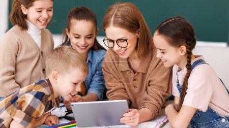 Glückliche Tutorin und optimistische Kinder, die lächeln und Videos auf dem Tablet ansehen, während sie sich während des Schulunterrichts um den Tisch versammeln