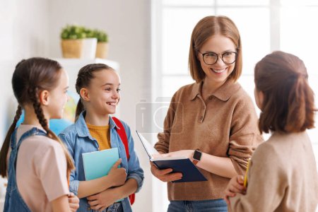 Photo for Happy woman teacher in casual clothes and glasses with book smiling and speaking with students while talking with pupils during literature lesson in sunlit classroom - Royalty Free Image