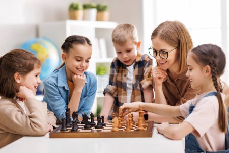 Photo for Happy female teacher in glasses pointing at board and explaining moves to cheerful children during chess lesson in daytime at school - Royalty Free Image