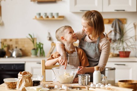 Téléchargez les photos : Famille joyeuse : maman et enfant garçon dans des tabliers faire de la pâtisserie à la table en bois dans la cuisine tout en cuisinant ensemble boulangerie - en image libre de droit