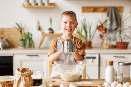 Foto de Sonriente niño lindo en delantal la adición de ingredientes en el tazón, mientras que la fabricación de masa para cocinar panadería en la cocina en casa - Imagen libre de derechos