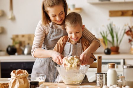 Foto de Familia alegre: mamá y niño en delantales haciendo pastelería en la mesa de madera en la cocina mientras cocinan panadería juntos - Imagen libre de derechos