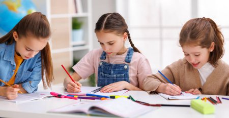 Photo for Positive little students girls     writing down data into notebook while sitting at table near classmates during test at school - Royalty Free Image