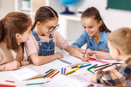 Photo for Smart girl with pigtails and glasses folding arms on table and smiling while communicating with classmates during lesson at school in daytime - Royalty Free Image