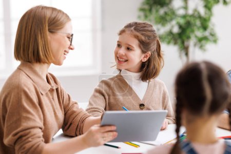 Photo for Happy female teacher in glasses smiling and looking at little student while sitting at table and showing data on tablet during lesson at school - Royalty Free Image