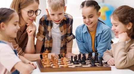 Photo for Happy female teacher in glasses pointing at board and explaining moves to cheerful children during chess lesson in daytime at school - Royalty Free Image