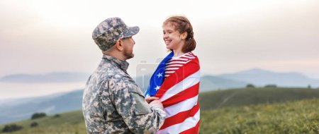 Foto de Feliz niña hija con bandera americana abrazando padre en uniforme militar regresó del ejército de los EE.UU., soldado masculino se reunió con la familia mientras estaba de pie en el prado verde en el día de verano - Imagen libre de derechos