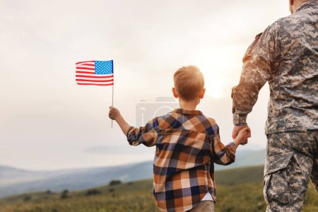 Photo for Rear view of military man father holding   son's hand   with american flag   and enjoying amazing summer nature view on sunny day, happy male soldier dad reunited with son after US army - Royalty Free Image