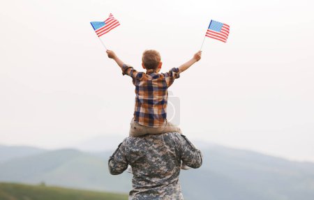 Foto de Vista trasera del padre militar llevando un pequeño hijo feliz con bandera americana en los hombros y disfrutando de la increíble vista de la naturaleza de verano en el día soleado, feliz padre soldado masculino se reunió con su hijo después del ejército de EE.UU. - Imagen libre de derechos