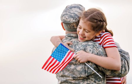 Photo for Happy little girl daughter with American flag hugging father in military uniform came back from US army, rear view of male soldier reunited with family while standing in green meadow on summer day - Royalty Free Image