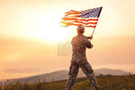 Photo for View from the back of a male soldier in the uniform of the American army waving the US flag on top of a mountain in a clearing at sunset - Royalty Free Image