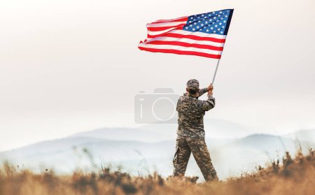View from the back of a male soldier in the uniform of the American army waving the US flag on top of a mountain in a clearing at sunset