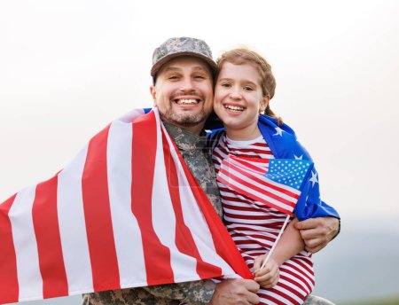 Téléchargez les photos : Bonne petite fille fille avec drapeau américain étreignant père en uniforme militaire est revenu de l'armée américaine, vue arrière du soldat masculin réuni avec la famille tout en se tenant debout dans la prairie verte le jour d'été - en image libre de droit