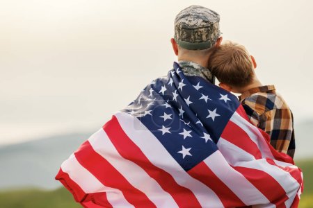 Happy little boy son with American flag hugging father in military uniform came back from US army, rear view of male soldier reunited with family while standing in green meadow on july 4th