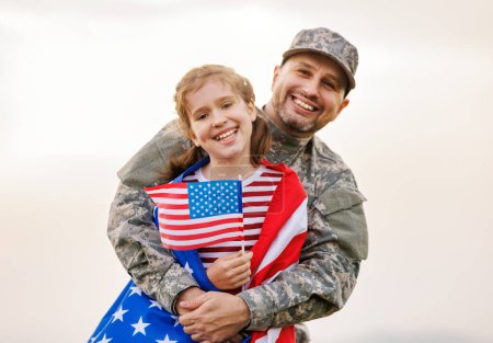 Téléchargez les photos : Bonne petite fille fille avec drapeau américain étreignant père en uniforme militaire est revenu de l'armée américaine, soldat masculin réuni avec la famille tout en se tenant dans la prairie verte le 4 juillet - en image libre de droit