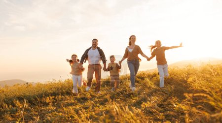 Téléchargez les photos : Heureux jeunes parents avec des enfants en vêtements décontractés courir avec la main tout en profitant du temps ensemble sur la prairie en soirée d'été - en image libre de droit