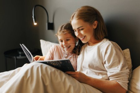 Photo for Cheerful family: woman and girl smiling and reading fairytale while relaxing on bed near lamp  at home - Royalty Free Image