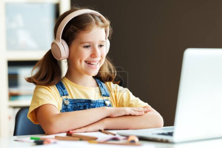 Photo for Positive child schoolgirl   with wireless headphones happy smiling, sitting at dusk and   looking at laptop screen during online lesson in daytime at home - Royalty Free Image