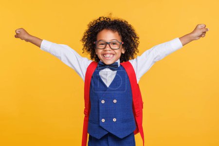 Téléchargez les photos : Garçon afro-américain excité en uniforme scolaire et lunettes levant le bras et souriant tout en célébrant le succès sur fond jaune - en image libre de droit