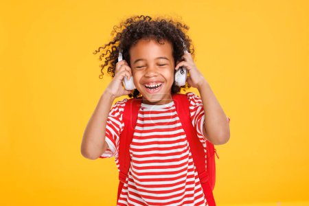 Téléchargez les photos : Africain américain riant garçon en t-shirt rayé ajuster les écouteurs tout en écoutant de la musique après les études scolaires sur fond jaune - en image libre de droit