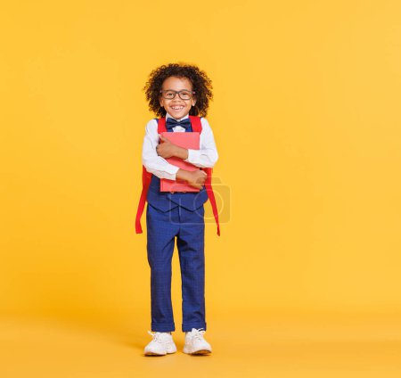 Foto de Cuerpo completo de niño afroamericano encantado en gafas abrazando libro y riendo en voz alta durante los estudios escolares contra el fondo amarillo - Imagen libre de derechos