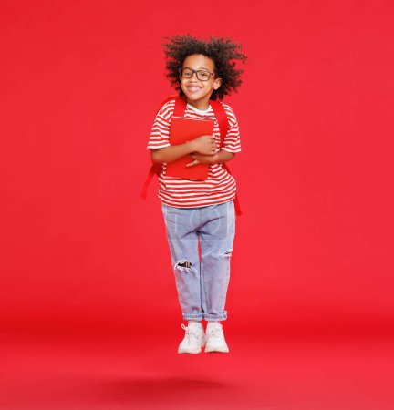 Téléchargez les photos : Garçon afro-américain pleine longueur en vêtements décontractés avec livre et saut sur fond rouge - en image libre de droit
