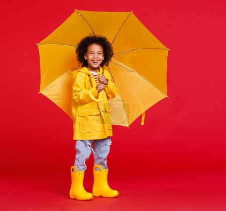 Téléchargez les photos : Drôle afro-américain garçon en imperméable et bottes avec parapluie debout tout en marchant sur fond rouge le jour de pluie - en image libre de droit