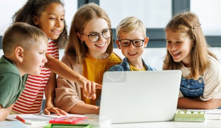 Photo for Happy female tutor and group of schoolchildren smiling and watching educational video on laptop during lesson in sunlit classroom in school - Royalty Free Image