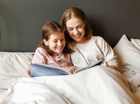 Foto de Familia alegre: mujer y niña sonriendo y leyendo cuento de hadas mientras se relaja en la cama cerca de la lámpara en casa - Imagen libre de derechos