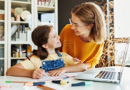 Photo for Smiling teacher or mother and schoolgirl browsing laptop while doing assignment together and studying remotely at home - Royalty Free Image