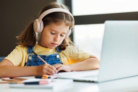 Photo for Focused child in wireless headphones sitting at desk and writing in planner near laptop during online lesson in daytime at home - Royalty Free Image
