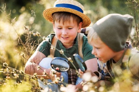 Foto de Dos niños pequeños con mochilas que buscan examinar plantas a través de lupa mientras exploran la naturaleza y el medio ambiente del bosque en un día soleado durante la lección de la escuela de ecología al aire libre - Imagen libre de derechos
