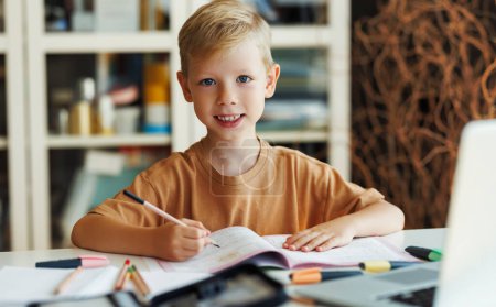 Foto de Niño enfocado en ropa casual sentado en el escritorio y escribiendo en copybook cerca de la computadora portátil durante la lección en línea durante el día en casa - Imagen libre de derechos