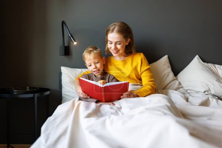 Téléchargez les photos : Famille joyeuse : femme et garçon fils souriant et lisant conte de fées tout en se relaxant sur le lit à la maison - en image libre de droit