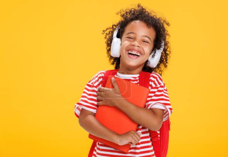 Foto de Niño risueño afroamericano con libro en camiseta a rayas ajustando auriculares mientras escucha la lección en línea después de la escuela estudia sobre fondo amarillo - Imagen libre de derechos