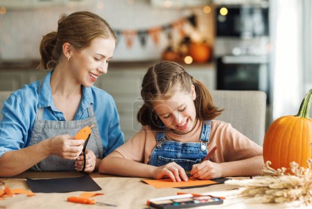 Photo for Happy smiling family mother and daughter making Halloween home decorations together while sitting at wooden table, mom and girl painting pumpkins and making paper cuttings - Royalty Free Image