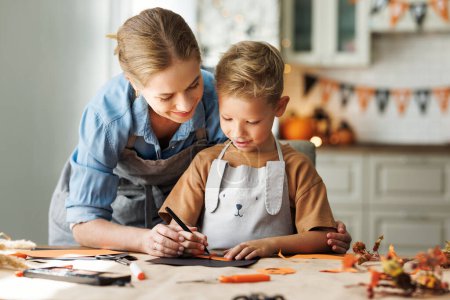 Photo for Happy smiling family mother and son making Halloween home decorations together while sitting at wooden table, mom and little boy painting pumpkins and making paper cuttings - Royalty Free Image