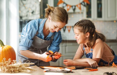 Photo for Happy smiling family mother and daughter making Halloween home decorations together while sitting at wooden table, mom and little girl painting pumpkins and making paper cuttings - Royalty Free Image