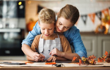 Photo for Happy smiling family mother and son making Halloween home decorations together while sitting at wooden table, mom and boy painting pumpkins and making paper cuttings - Royalty Free Image
