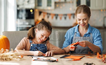 Photo for Happy smiling family mother and daughter making Halloween home decorations together while sitting at wooden table, mom and girl painting pumpkins and making paper cuttings - Royalty Free Image