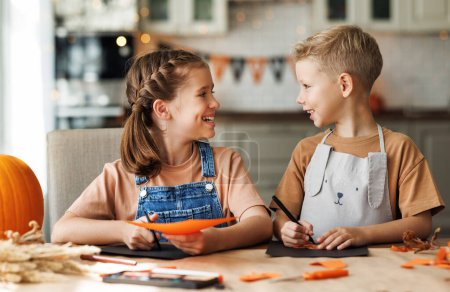 Photo for Portrait of happy siblings kids boy and girl making Halloween home decorations together while sitting at wooden table, brother and sister painting pumpkins and making paper cuttings - Royalty Free Image