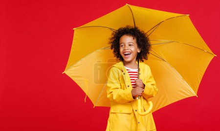 Foto de Alegre chico afroamericano en impermeable riendo y llevando paraguas amarillo en el hombro en día lluvioso sobre fondo rojo - Imagen libre de derechos