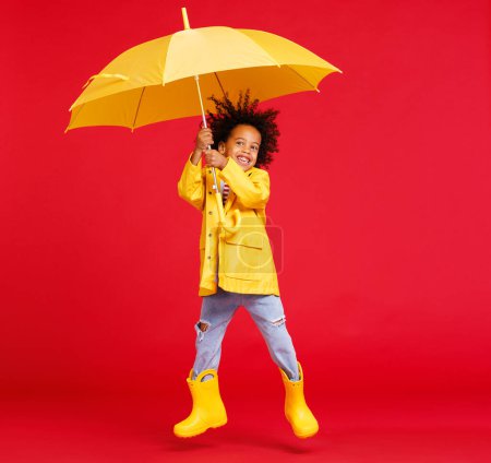 Téléchargez les photos : Drôle afro-américain garçon en imperméable et bottes avec parapluie saut tout en marchant sur fond rouge le jour de pluie - en image libre de droit