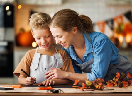 Téléchargez les photos : Happy souriant famille mère et fils faisant Halloween décoration de maison ensemble tout en étant assis à la table en bois, maman et garçon peinture citrouilles et faire des découpes de papier - en image libre de droit