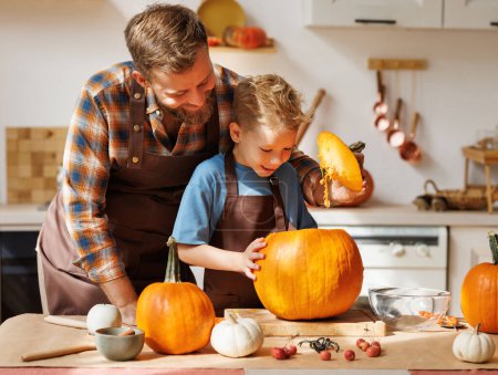 Foto de Lindo niño feliz ayudando a su padre a tallar calabaza de Halloween mientras está de pie en la cocina en casa y preparándose para las vacaciones de otoño, hijo de familia y papá haciendo Jack-o-Lantern juntos - Imagen libre de derechos